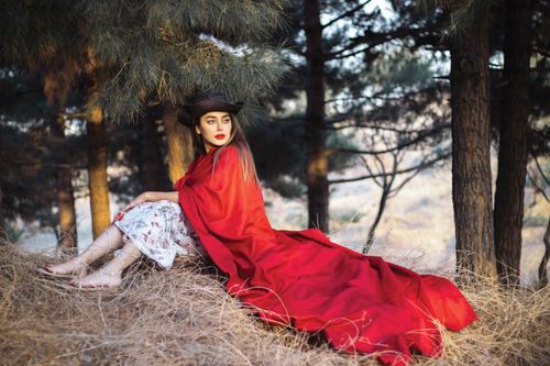 Free A beautiful woman in a red cape and hat sitting in a serene forest setting. Stock Photo