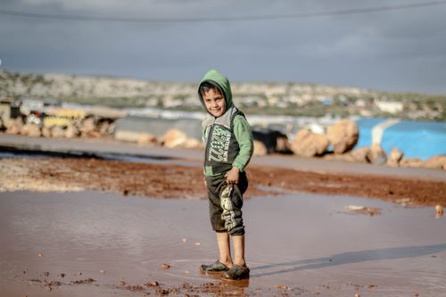 Free A young boy joyfully stands in a muddy puddle in a refugee camp in Idlib, Syria, capturing a moment of innocence. Stock Photo