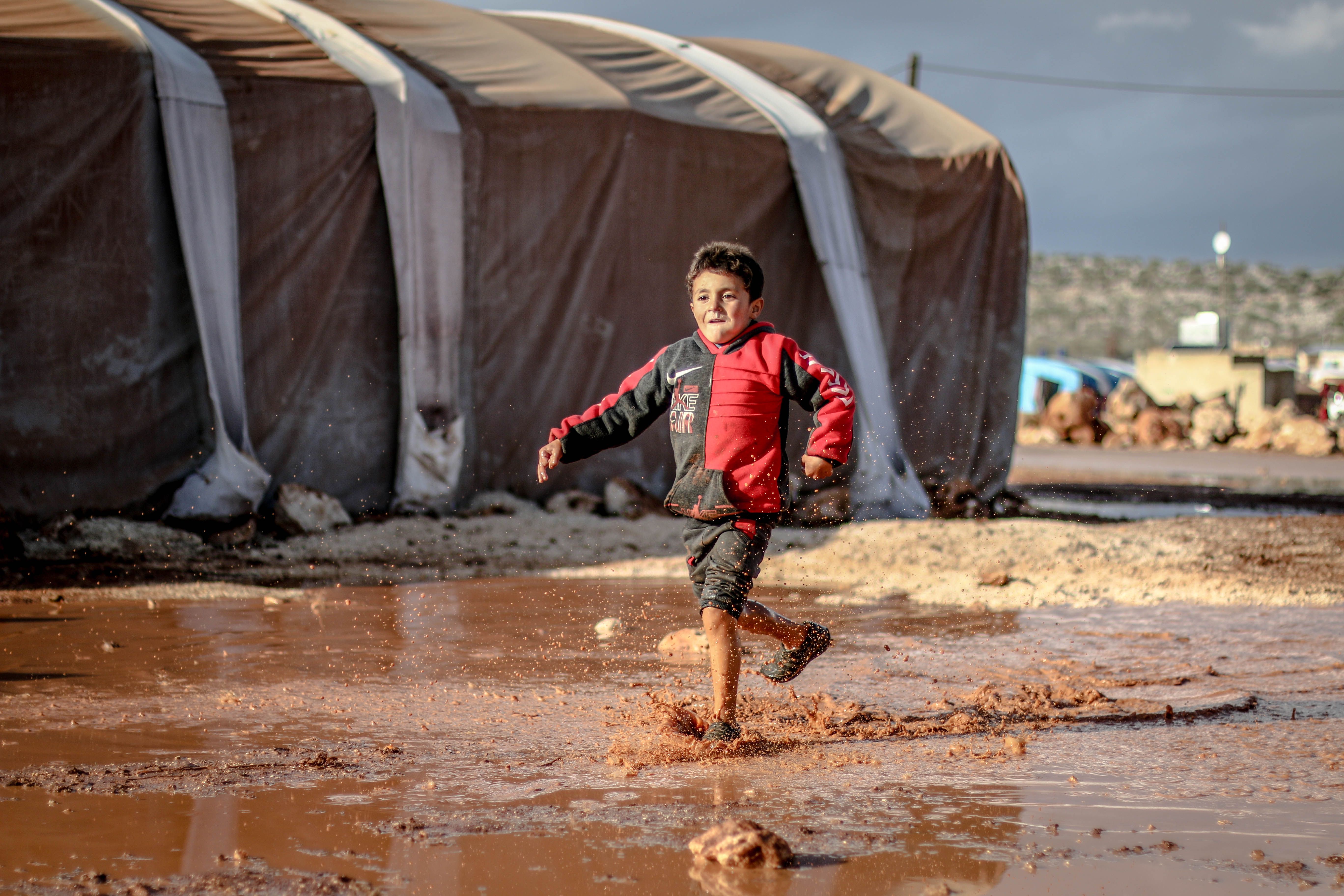 A joyful young boy runs through a muddy puddle in a refugee camp in Idlib, Syria, encapsulating resilience and innocence.