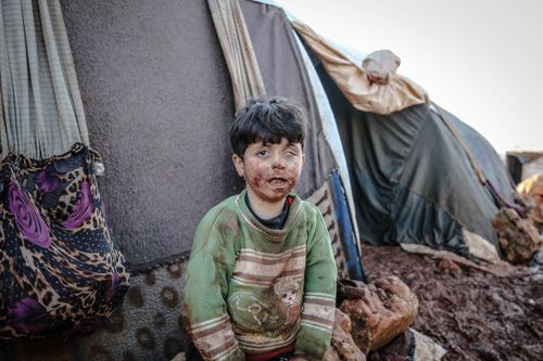 Free Young child covered in mud sits near tents in a refugee camp in Idlib, Syria. Stock Photo