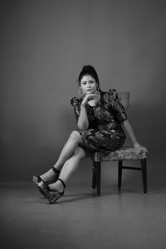 A thoughtful woman in a floral dress sitting on a chair in a monochrome studio portrait, exuding elegance.