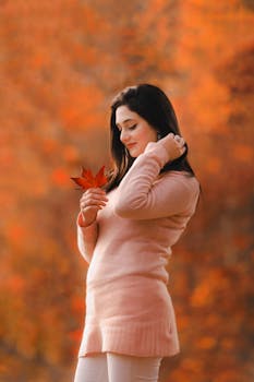 Woman in pink sweater poses with a maple leaf in an autumn setting, embracing the seasonal warmth.