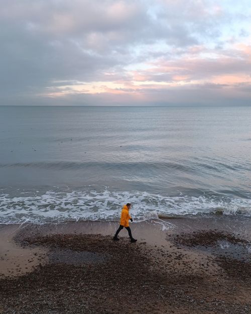 Free A person in warm clothing walks along a cloudy winter beach with calm waves. Stock Photo