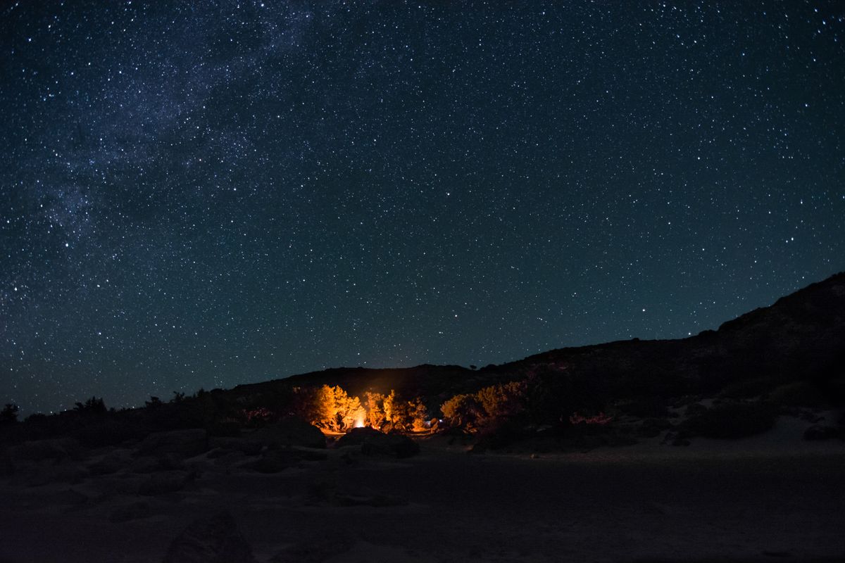 A small campfire glowing at a backcountry campsite beneath a mountain and starry night sky