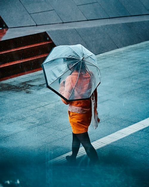 Free Woman in orange dress holding a transparent umbrella, walking down a rainy street. Stock Photo