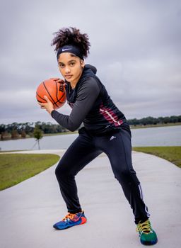 Confident young woman in activewear playing basketball on a cloudy day by a lake.