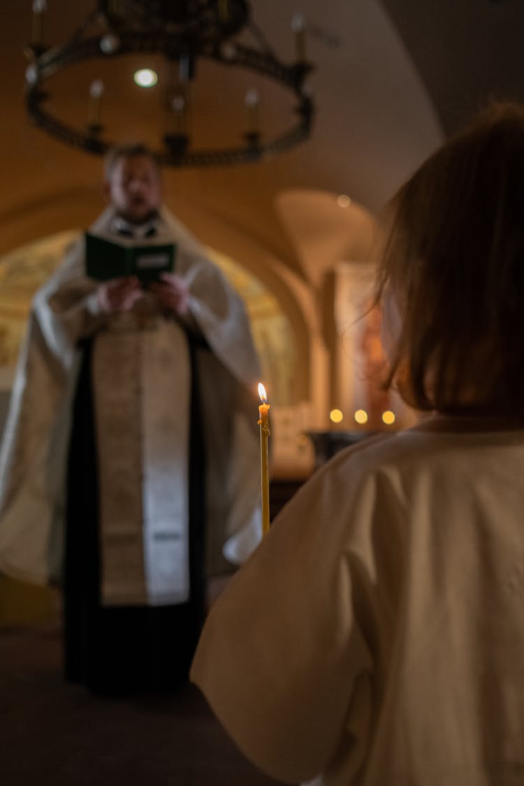 A Kid Holding A Candle While Listening To A Bishop Talking In Front Of Her