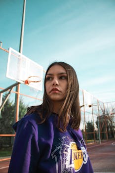 Portrait of a woman in a Lakers hoodie on an outdoor basketball court under a clear sky.