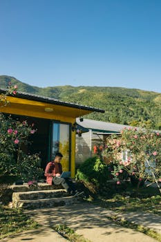 Man sitting outside a modern house with a laptop surrounded by nature and flowers.