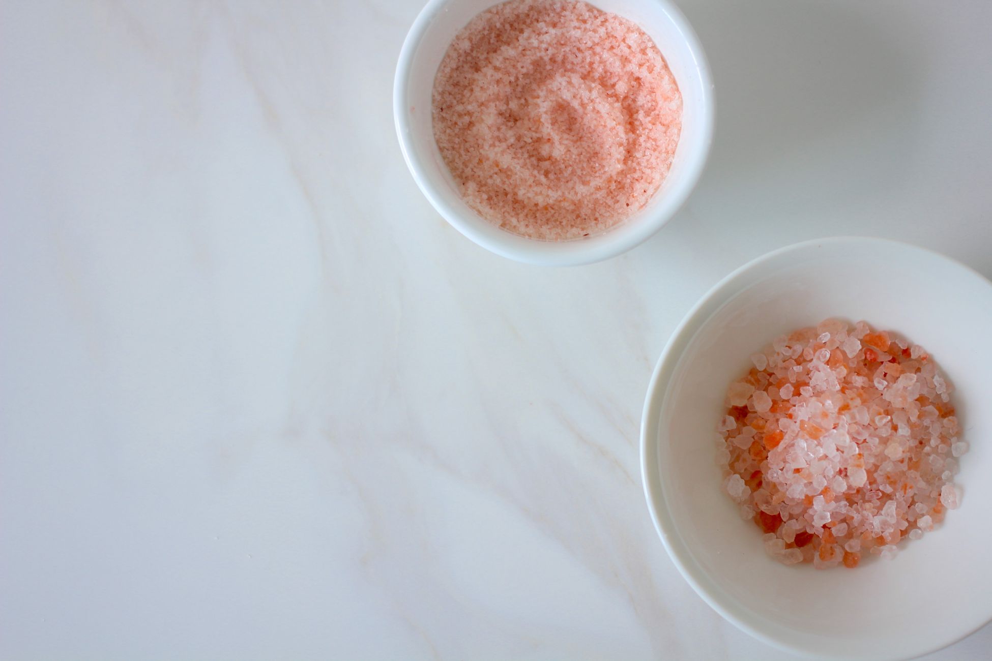 Two white ceramic bowls containing different textures of pink Himalayan salt on a marble surface.