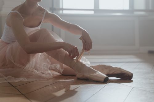 Free Ballerina sitting on wooden floor tying pointe shoes, preparing for ballet practice in a serene studio setting. Stock Photo