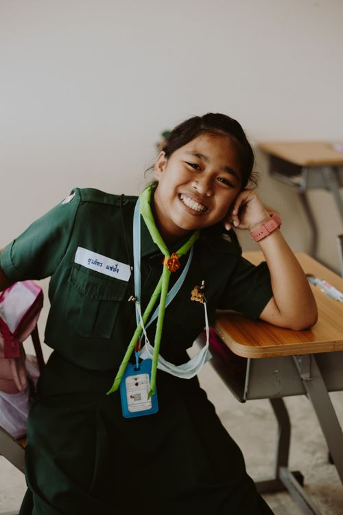 Free Cheerful Asian student in uniform sitting at a wooden desk indoors. Stock Photo