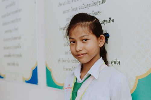 Free Portrait of a young Thai schoolgirl in uniform smiling against an educational poster background. Stock Photo