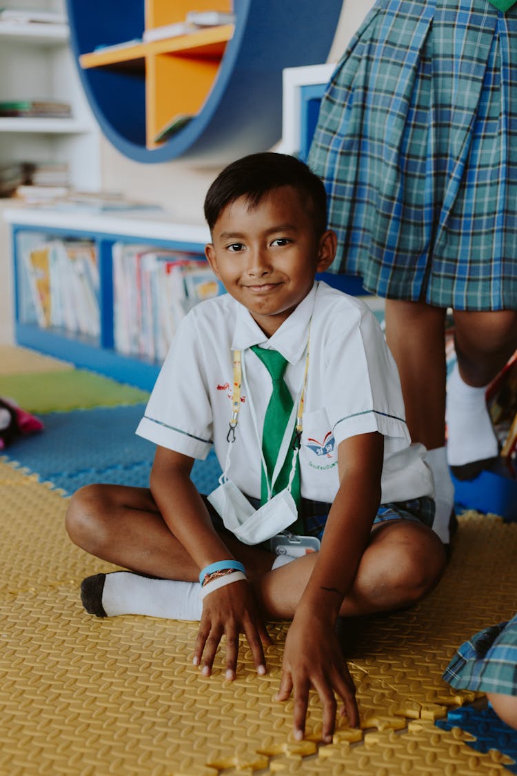 Portrait Of Smiling Boy In School Uniform Sitting On Floor In Classroom