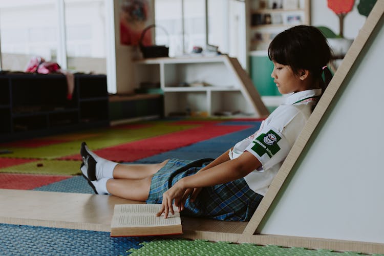 Girl In School Uniform Reading Book On Floor In Classroom