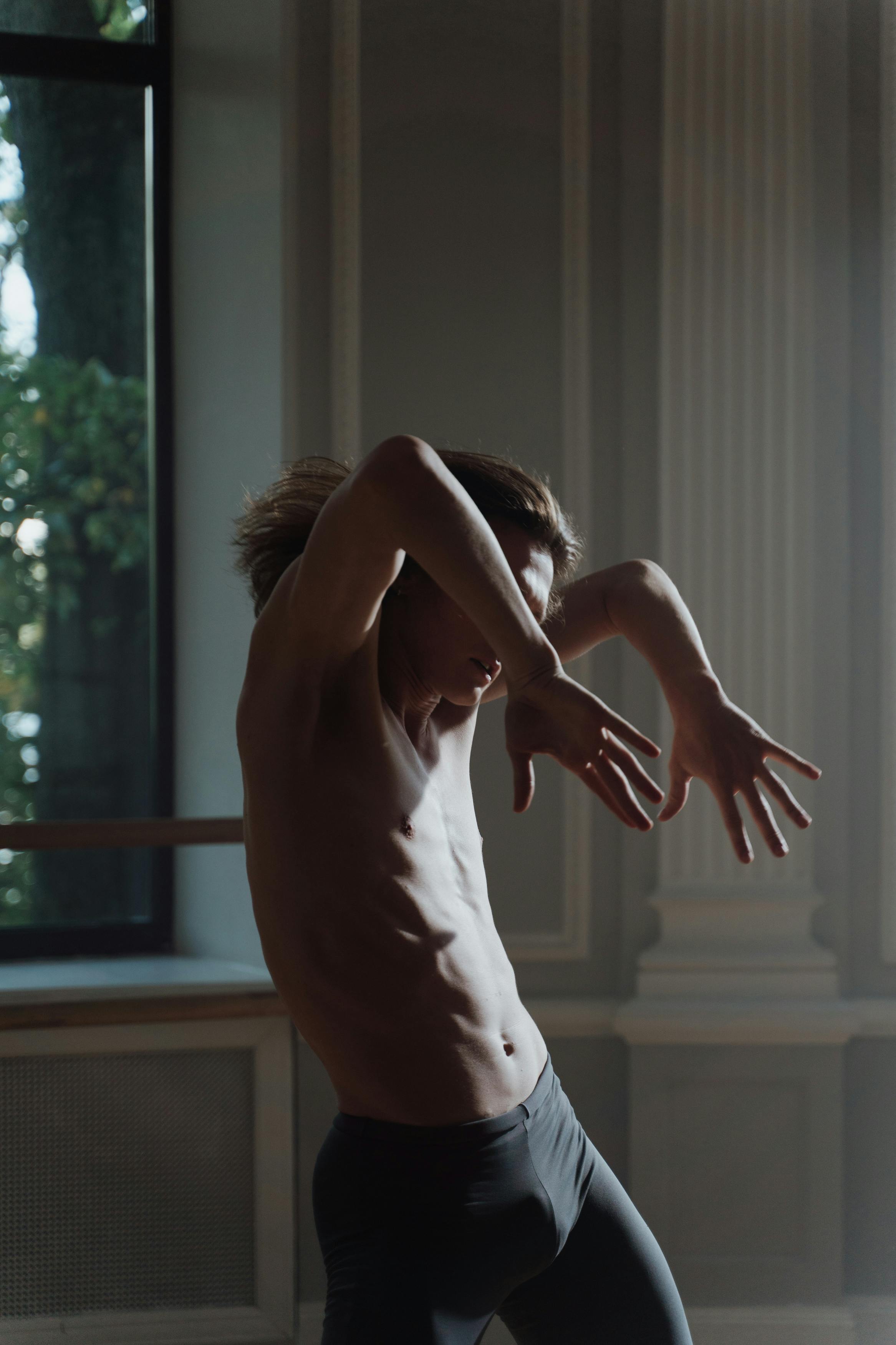 A shirtless man performs an expressive dance pose in a sunlit studio, embodying grace and strength.