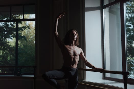 A shirtless male dancer gracefully performs a ballet pose by a window in a dimly lit studio.
