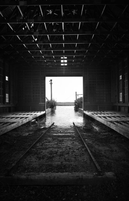 Free Black and white photo of an empty boathouse interior with train tracks leading to a lakeshore. Stock Photo