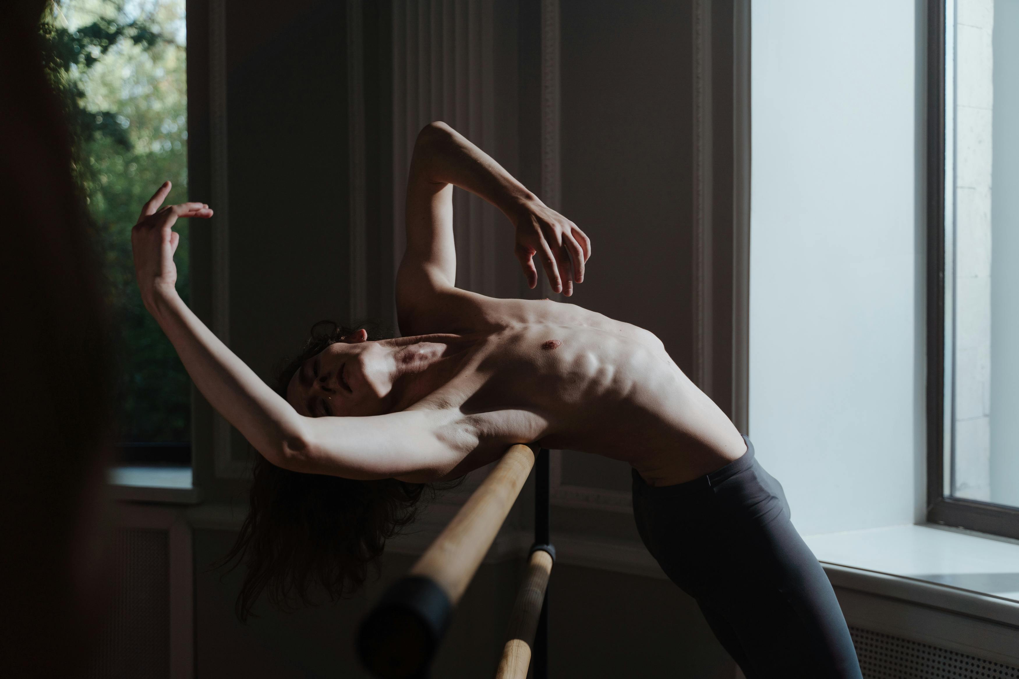 A shirtless male dancer showcases flexibility and strength in an expressive pose at the barre.