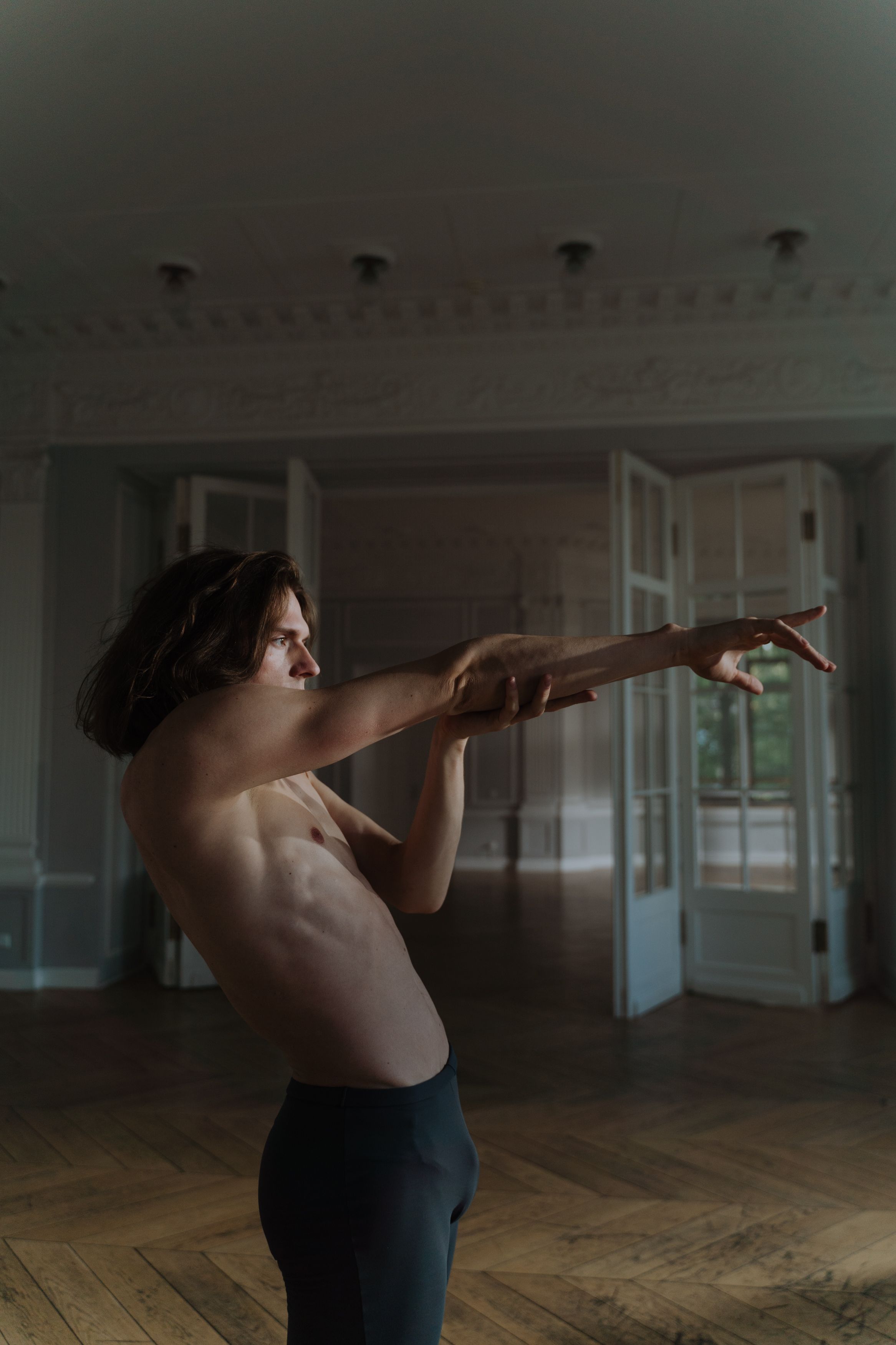 Shirtless male dancer performing a contemporary dance in a sunlit elegant room, showcasing flexibility and grace.