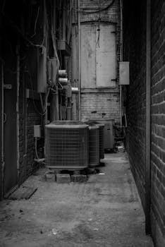 Black and white photo of urban alley featuring air conditioning units, cables, and walls.