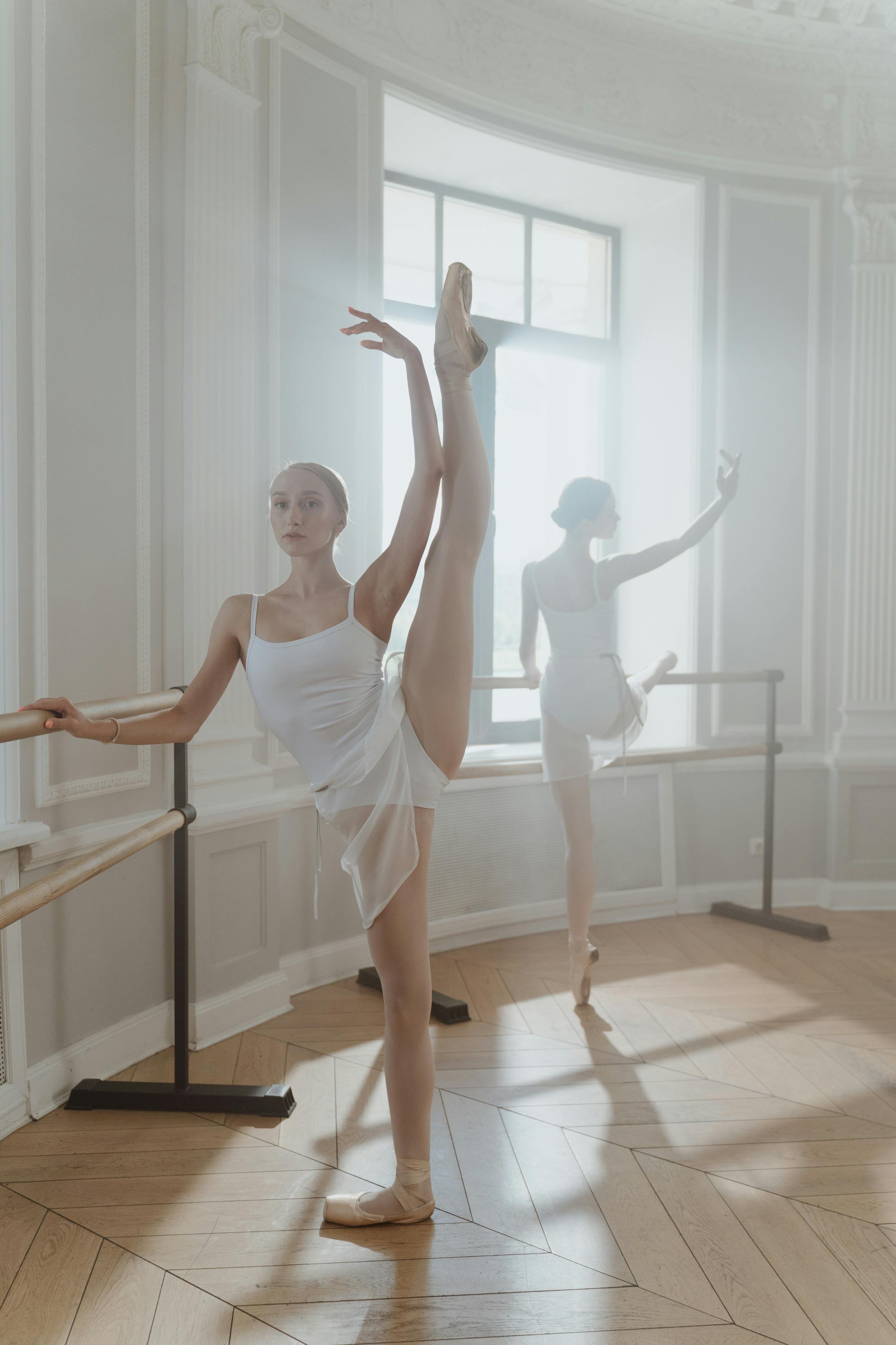 ballerina in white tank top standing with leg up on wooden floor in front of ballerina beside window