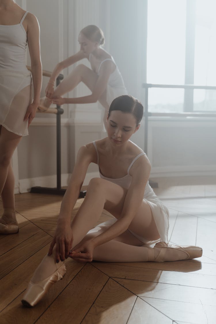 A Ballet Dancer Fixing Her Pointe Shoes On A Wooden Floor