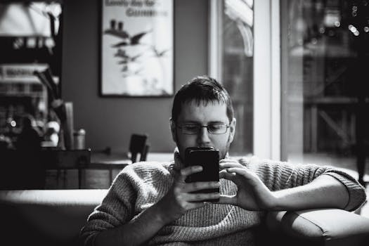 A man in a cozy indoor space engrossed with his smartphone, sipping coffee.
