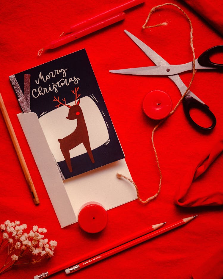 Christmas Card, Scissors, Candles And Pencils On Red Textile