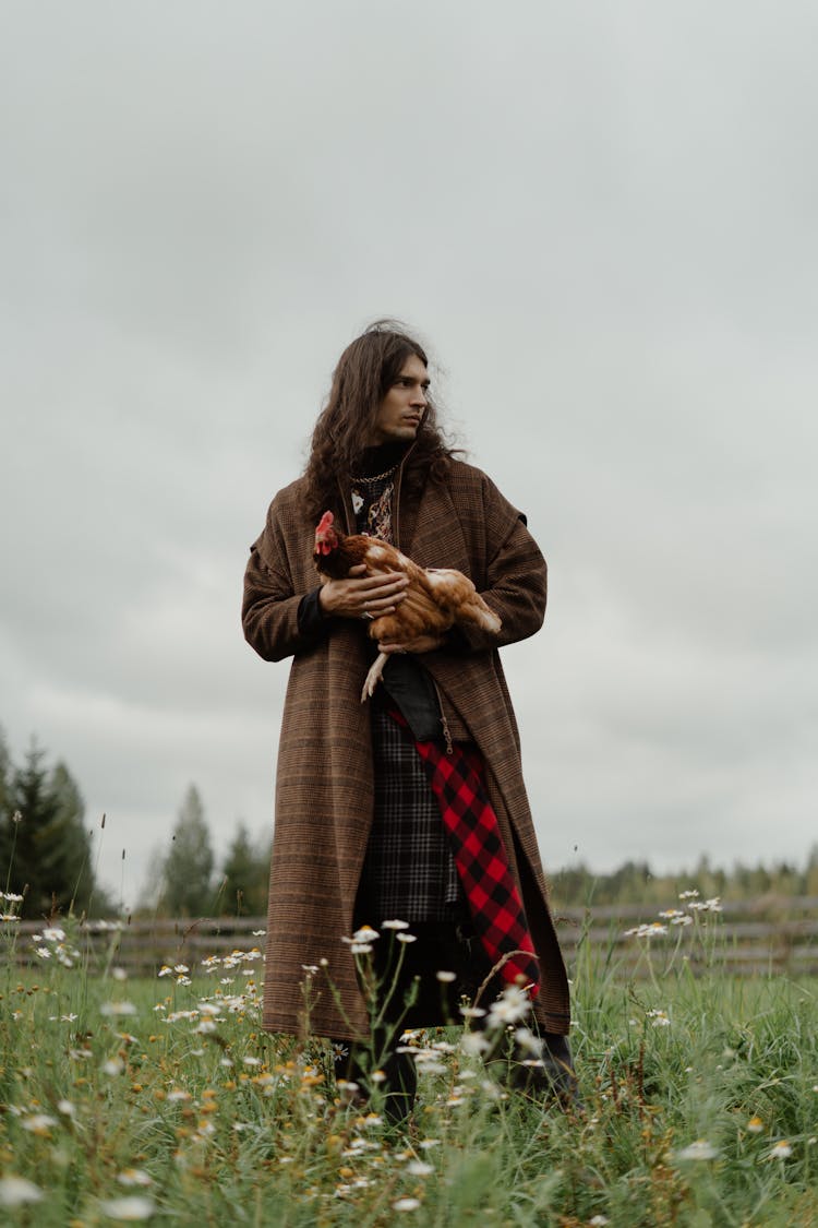 A Man In Brown Coat Standing On Green Grass While Carrying A Rooster