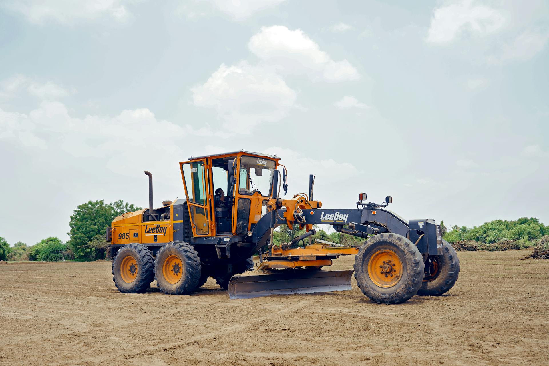 A motor grader working in an open field under a cloudy sky at Sanand, India.