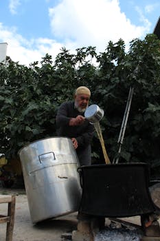 Elderly man pouring liquid into a large pot outdoors, near fig trees, under a blue sky.