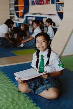 A young girl in uniform reading a book while sitting cross-legged in a colorful classroom.