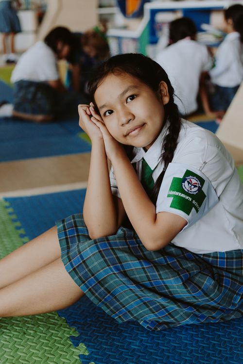 Free Smiling Thai girl in school uniform sitting on a colorful classroom floor, looking at the camera. Stock Photo