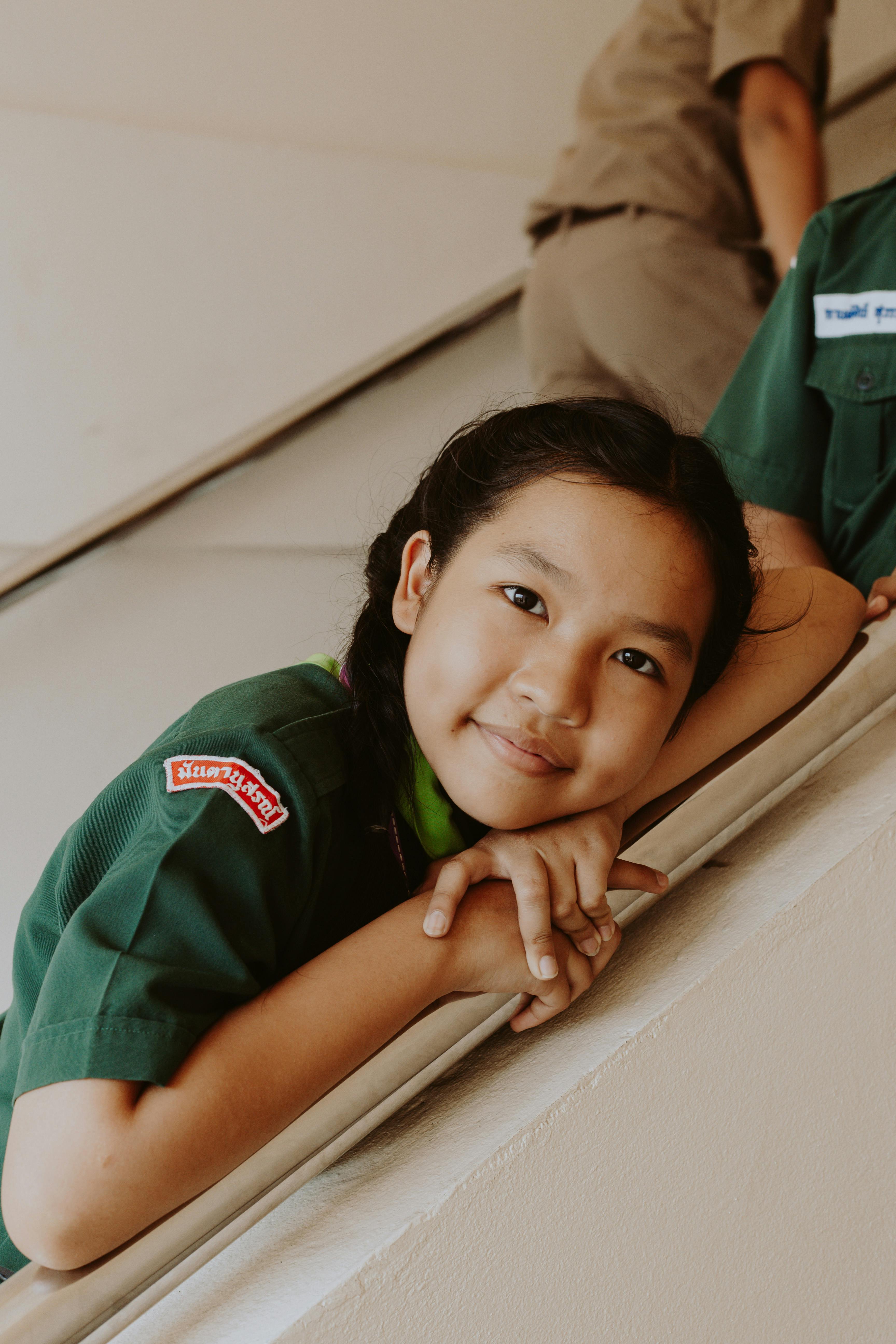 A girl in a green scout uniform smiling while resting on a railing indoors.