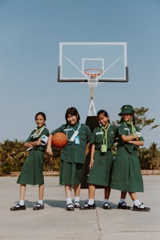 Smiling schoolgirls in green uniforms posing on an outdoor basketball court under a clear sky.