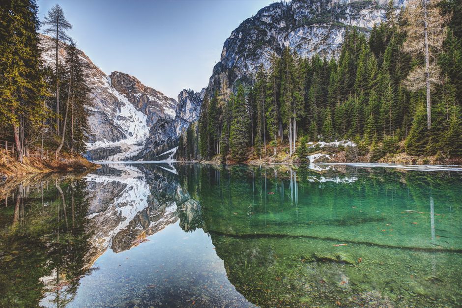 Tranquil lake reflecting snowy mountains and evergreen trees in a serene landscape.