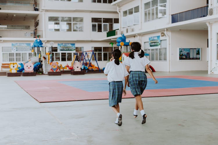 Rear View Of Two Girls In School Uniforms Running In School Gymnasium