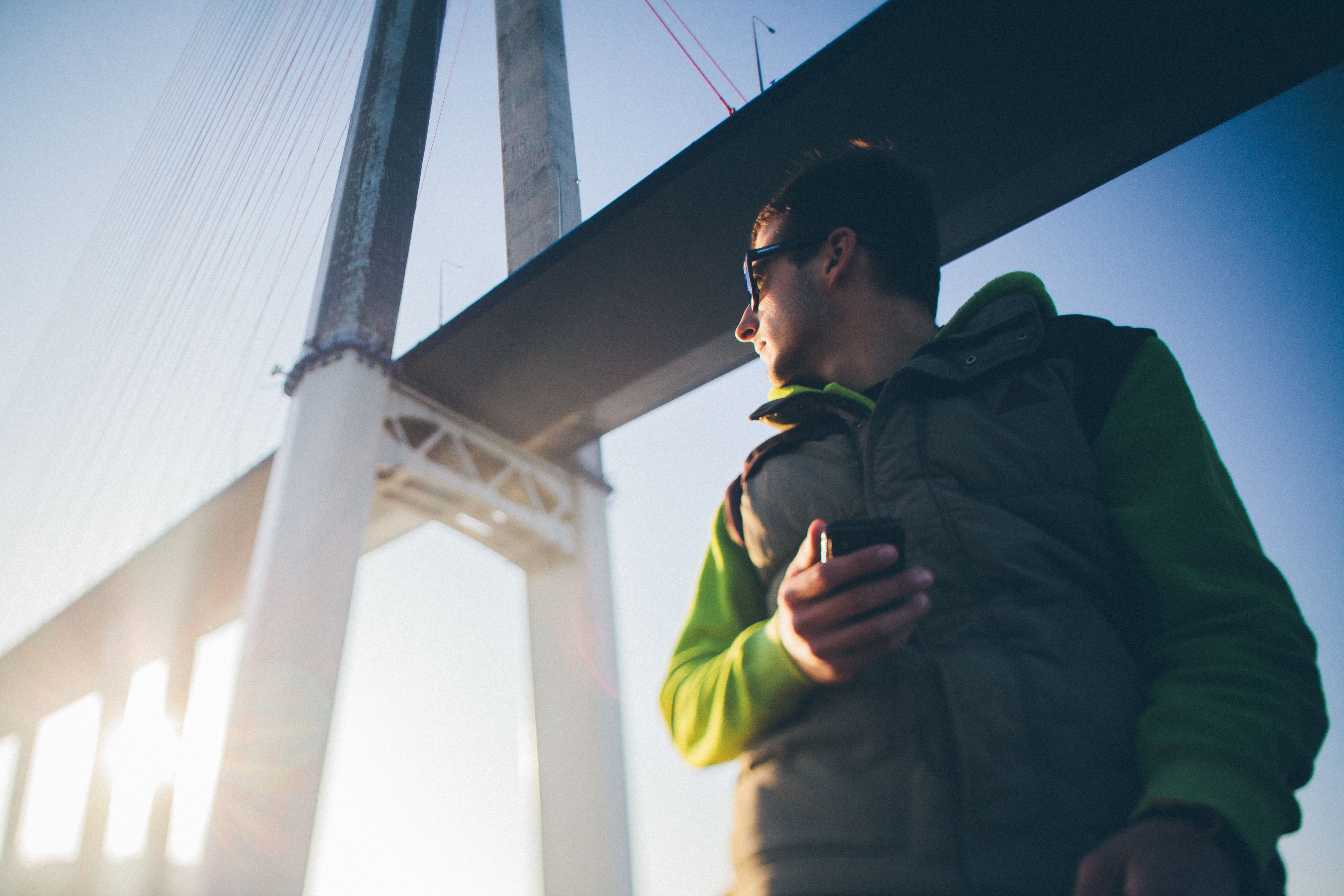 A young man in sunglasses holding a phone beneath a large bridge, enjoying a sunny day.