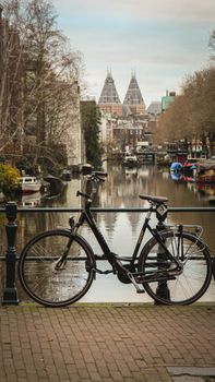 Black bicycle parked by a scenic canal in Amsterdam with city buildings and reflection in the water.