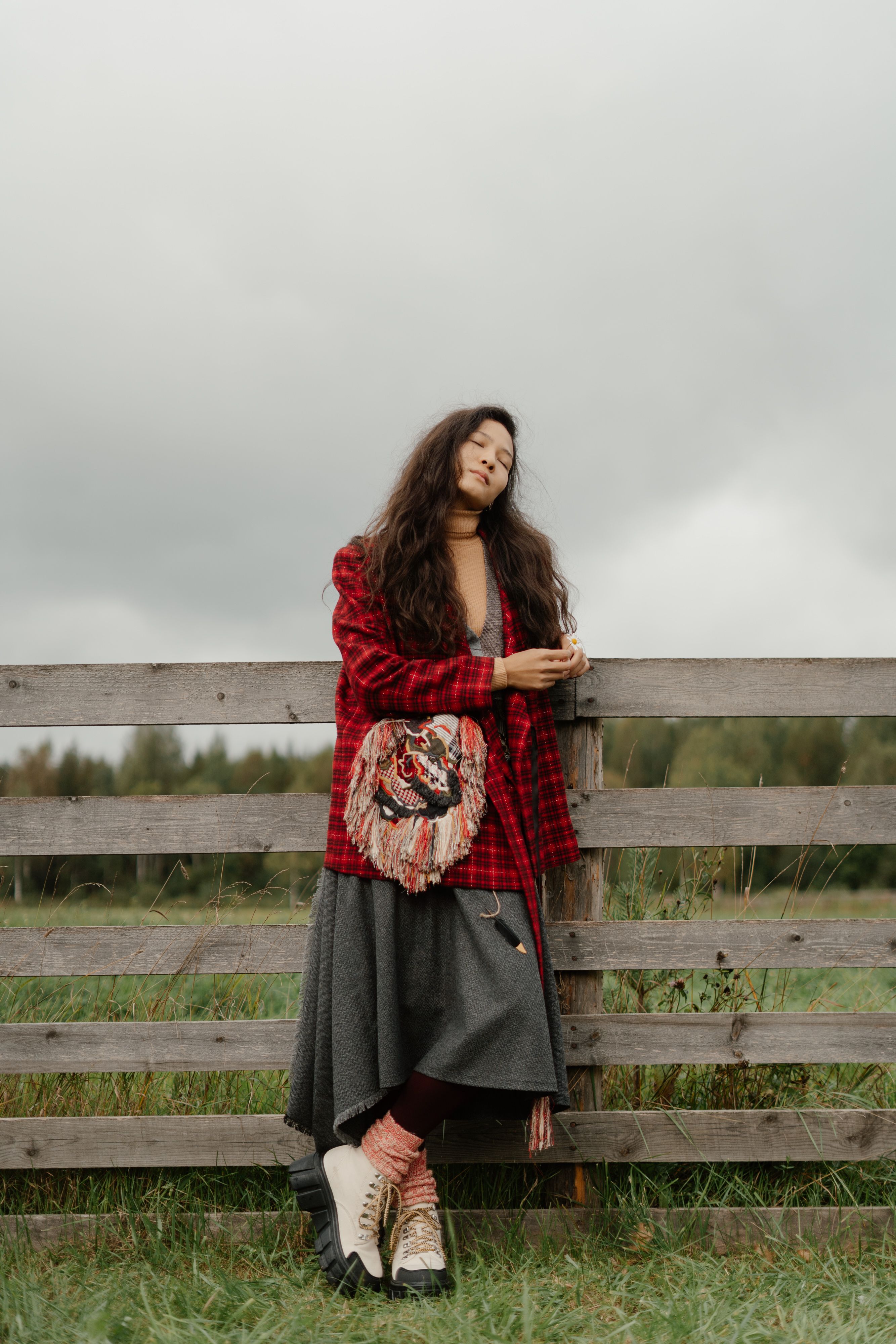 A young woman in a red jacket leans against a wooden fence on an overcast day, expressing tranquility.