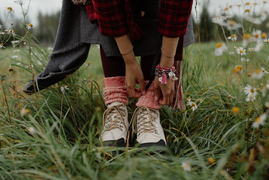 A woman in a plaid jacket ties her hiking boots surrounded by wild daisies in a meadow.