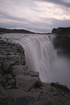 A dramatic waterfall with moody skies cascading over rugged rocks in a breathtaking natural setting.