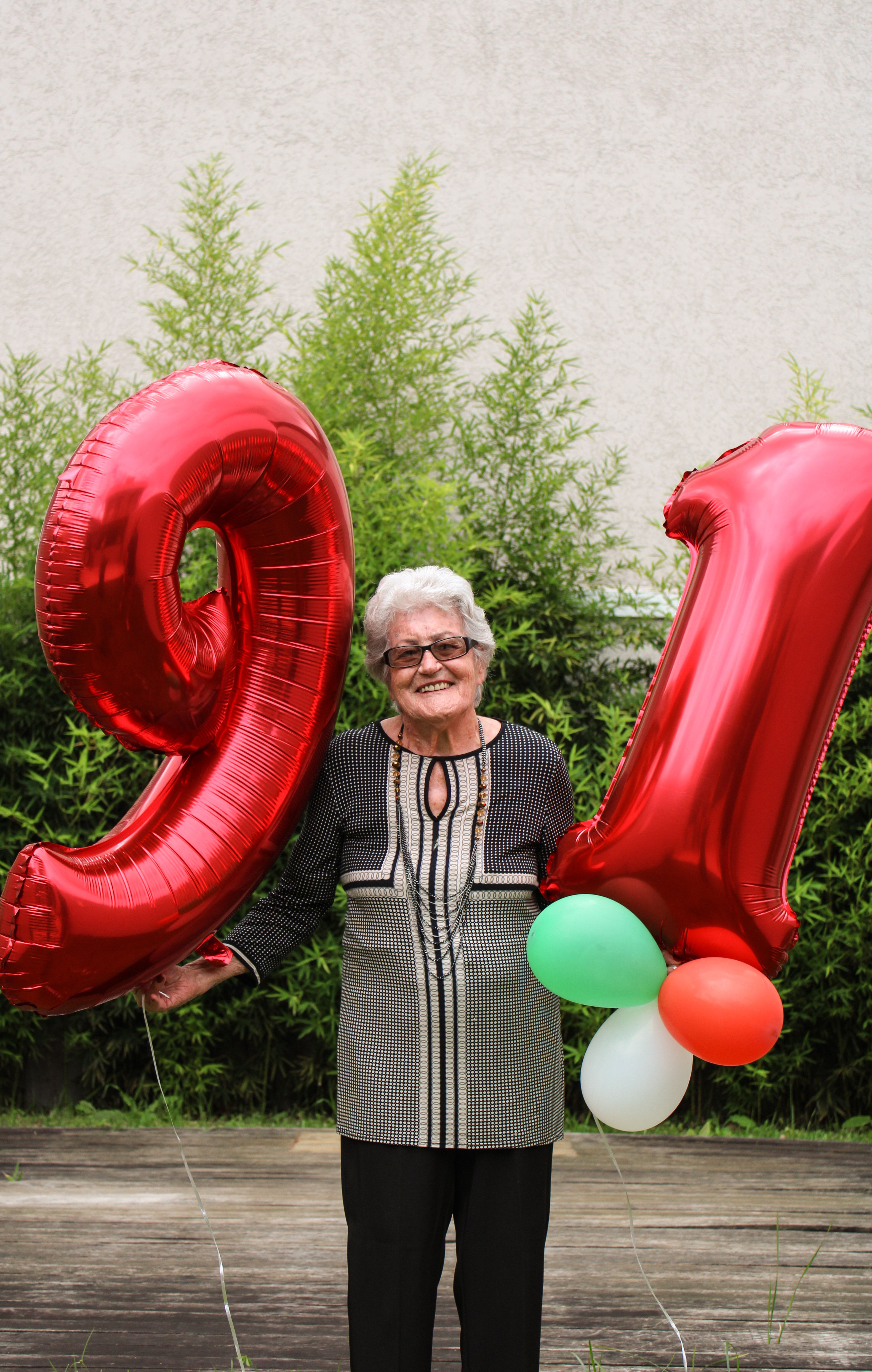 Elderly woman smiling while holding 91 balloons in a garden setting.