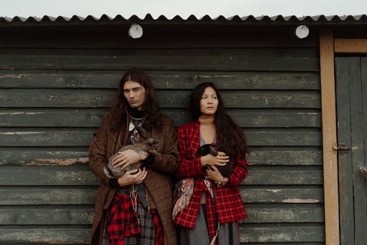 A bohemian couple holding rabbits in front of a rustic wooden barn, showcasing a unique fashion style.