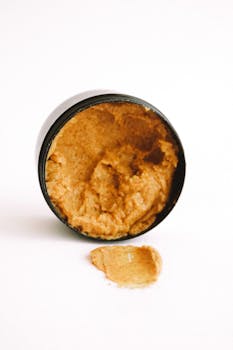 Open jar of natural body scrub with some spilled scrub against a white background.