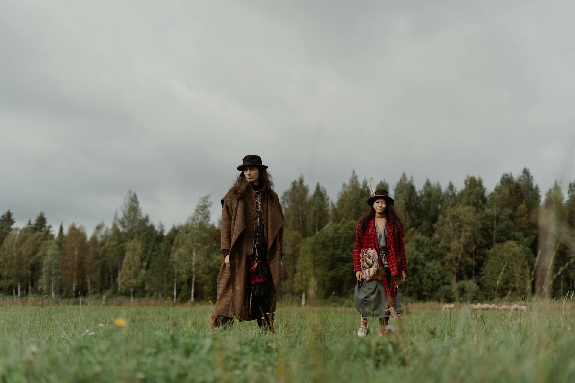 Deux adultes dans des manteaux et des fedoras à la mode marchent dans une campagne herbeuse, entourés d'arbres.