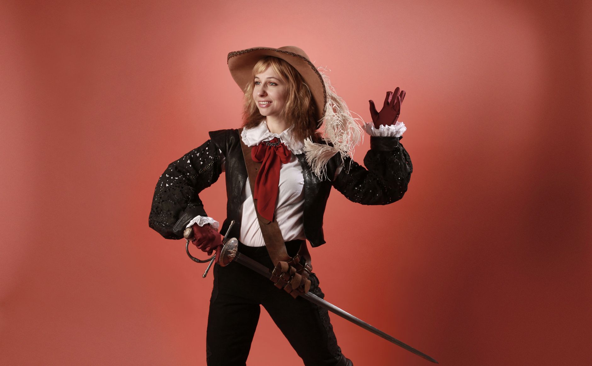 Studio portrait of a woman in a musketeer costume holding a rapier against a red backdrop.