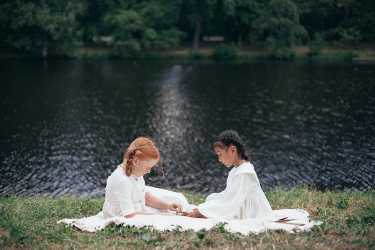 Two Girls In White Dresses Sitting On Riverbank