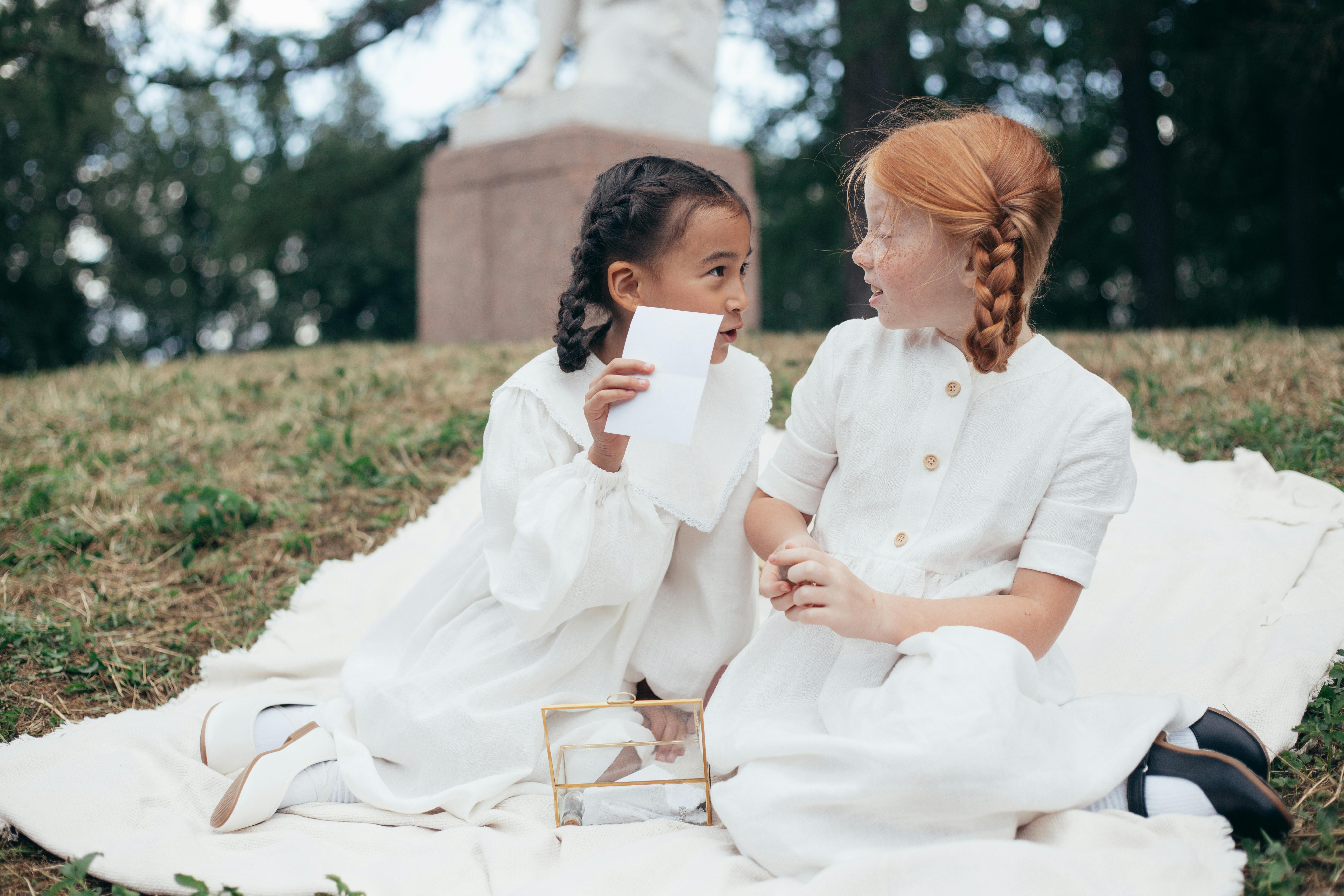 Two young girls sharing a secret while sitting on a blanket in a sunny park.